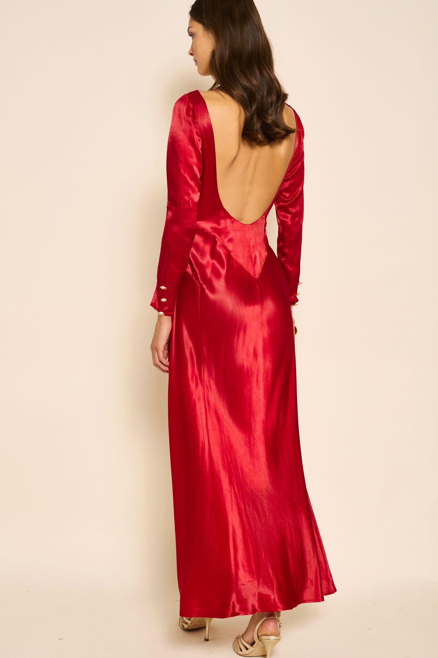 A woman with wavy brown hair models the Devina - Crimson, a shiny red satin evening dress with long sleeves and a deep open back. Facing away and glancing to the side, she finishes her look with gold high-heeled sandals.