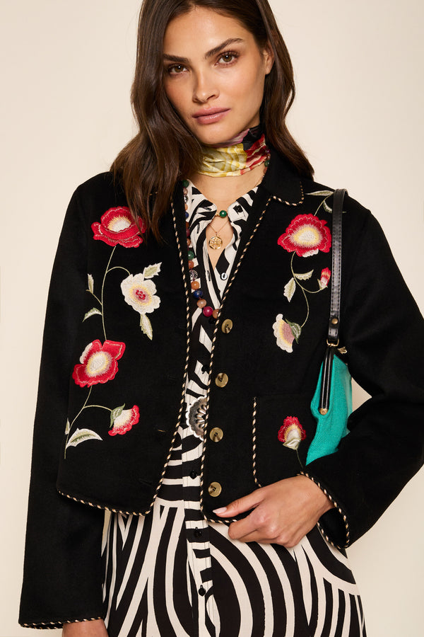 A woman wears the Sunday - Poppy Embroidery Black jacket over a zebra print dress, styled with wavy brown hair, a patterned scarf, and a turquoise bag.