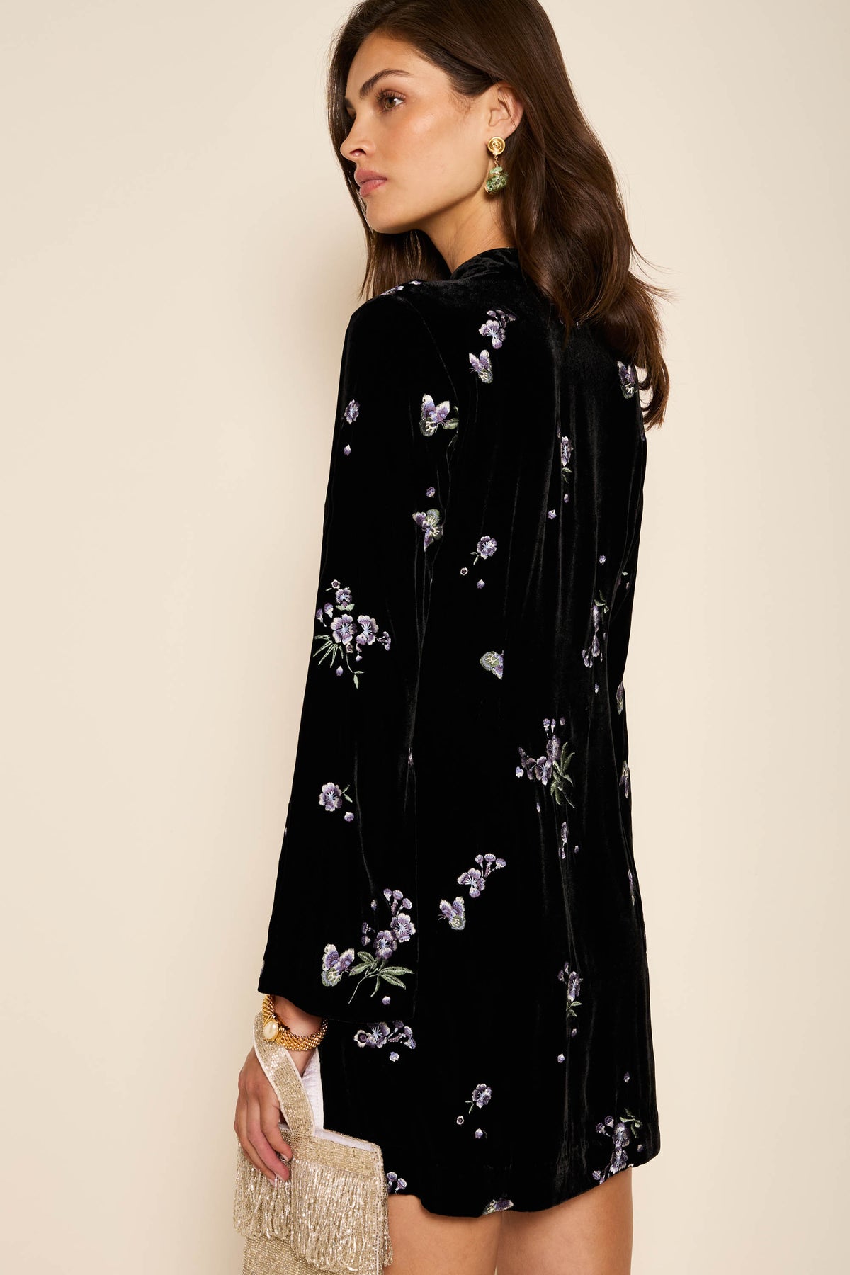 A woman in the Calla - Blossom Embroidery Blue dress, featuring blue floral embroidery, holds a cream fringed clutch. She has wavy brown hair, green statement earrings, and stands against a beige background.