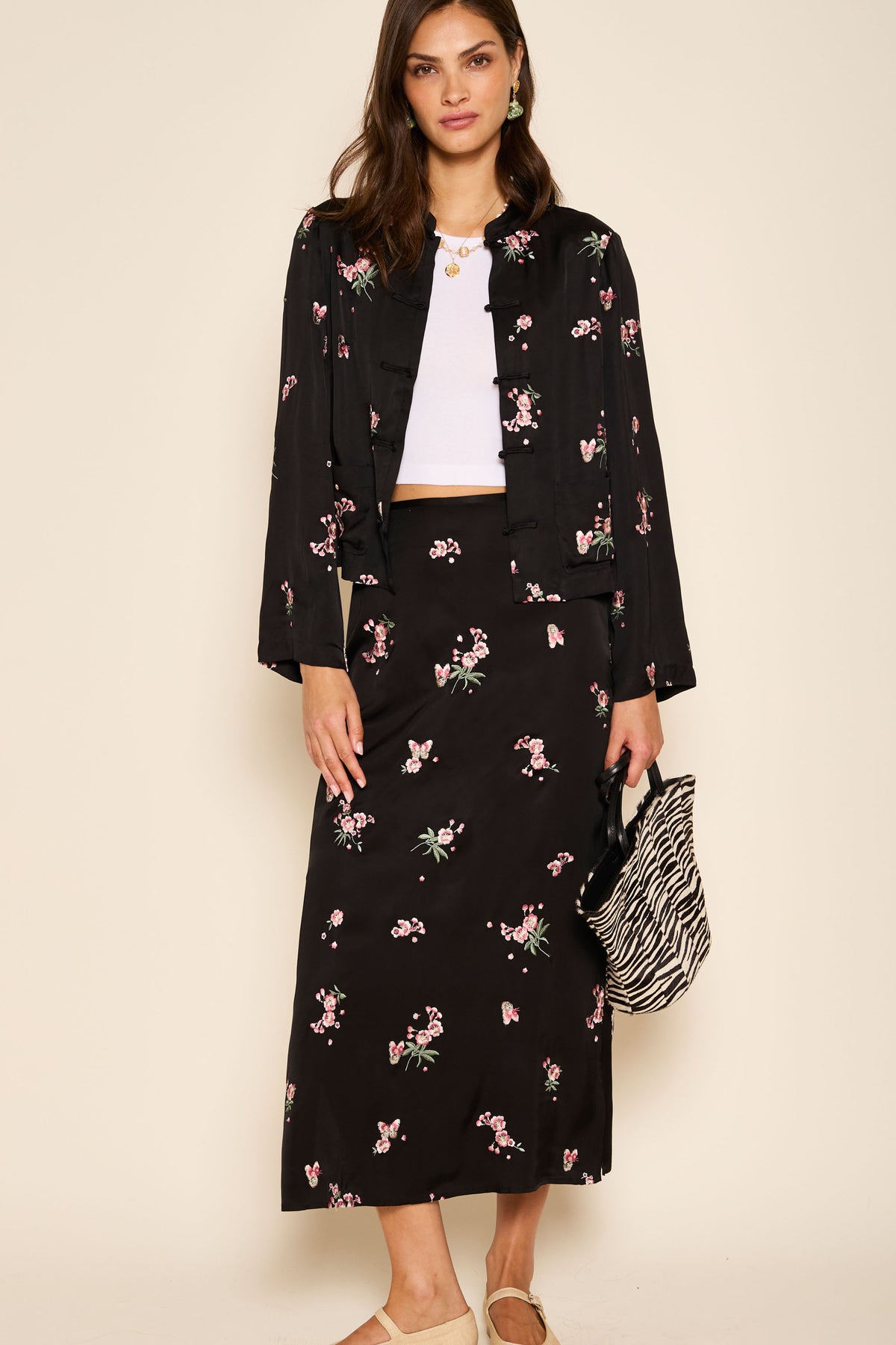 A woman stands against a beige background in the Indiya - Blossom Embroidery Pink column skirt, paired with a black floral jacket, white crop top, beige flats, and zebra-print bag—available in both regular and petite sizes.