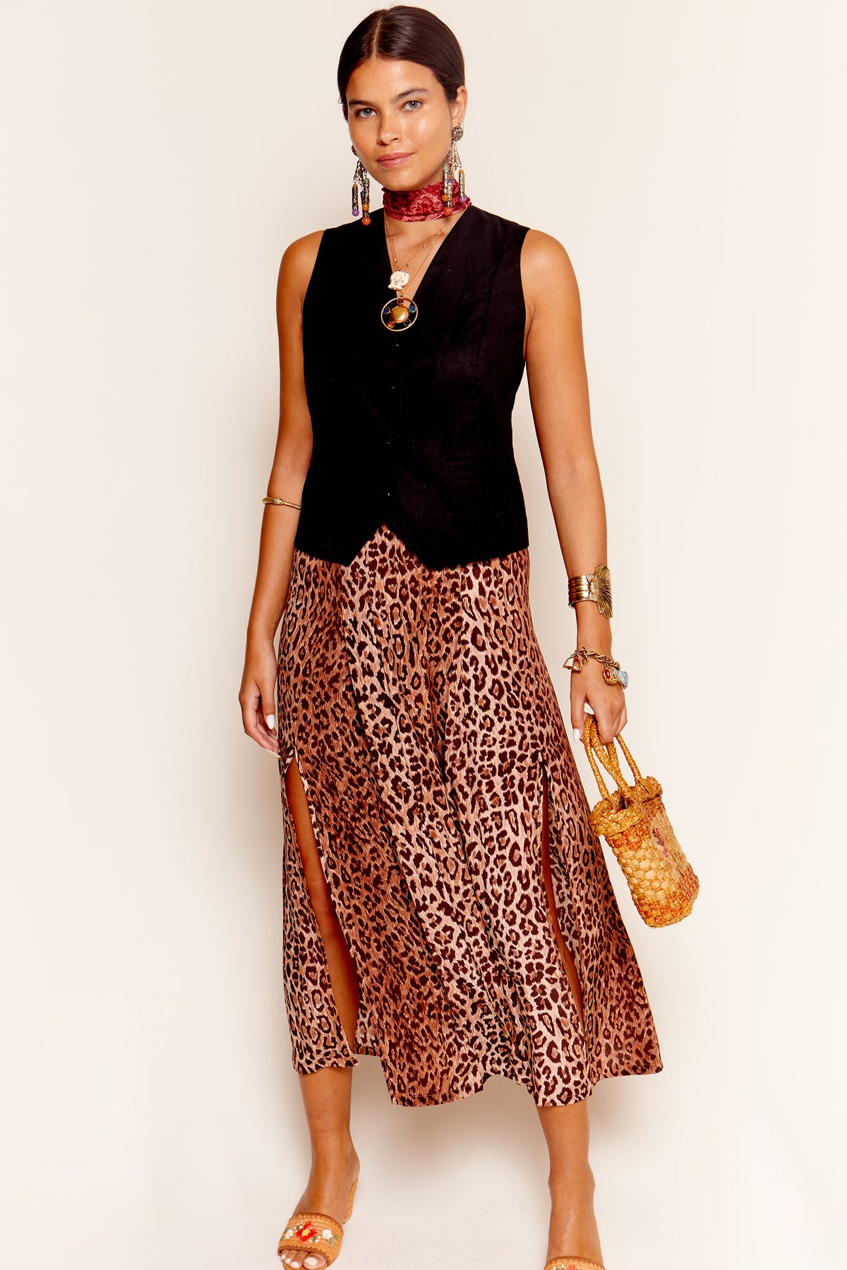 A woman poses against a light background wearing the Georgia - Leopard silk midi skirt with side slits, a black sleeveless vest, gold sandals, statement jewelry, and holding a small woven bag.