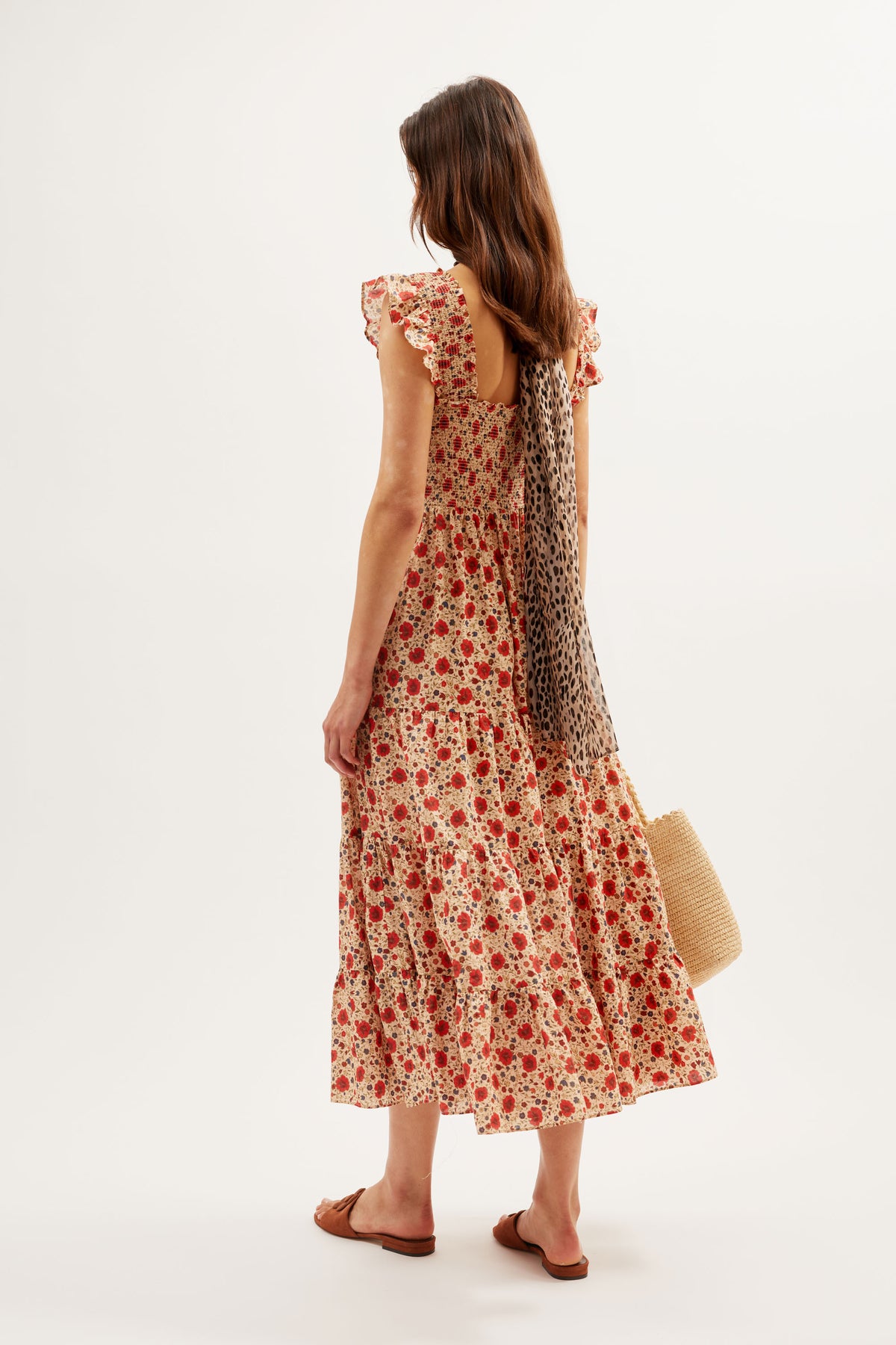 A woman faces away wearing the Kendall – Eva Floral Cream sleeveless, mid-length dress with ruffled shoulders, brown sandals, a straw bag, and a spotted scarf tied at the back. The background is white.