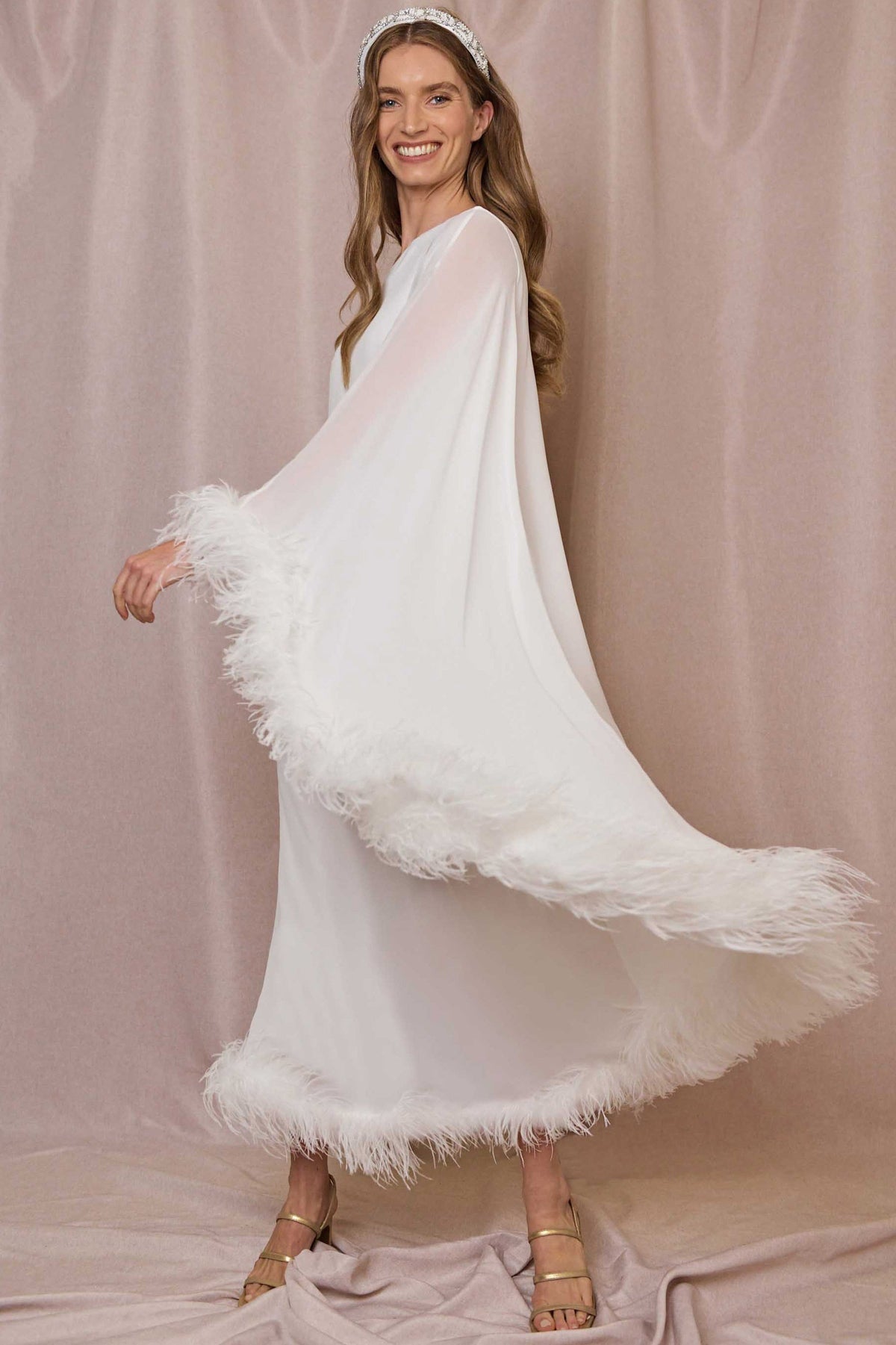A smiling woman wears the Liza - Ivory Feathers dress, featuring feather-trimmed georgette with flowing ivory hem and sleeves, gold sandals, and a jeweled headband against a soft pink background for an elegant look.