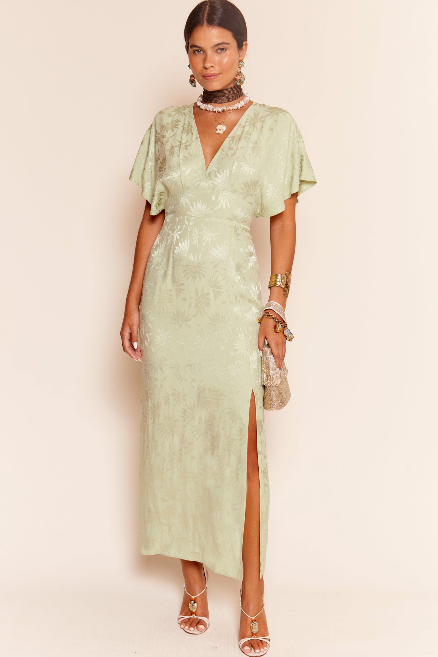 A woman wears the Sadie - Daisy Jacquard Sage dress, featuring thirties-inspired kimono sleeves, a deep V-neck, and side slit. She accessorizes with layered necklaces, bangles, earrings, and a small purse.