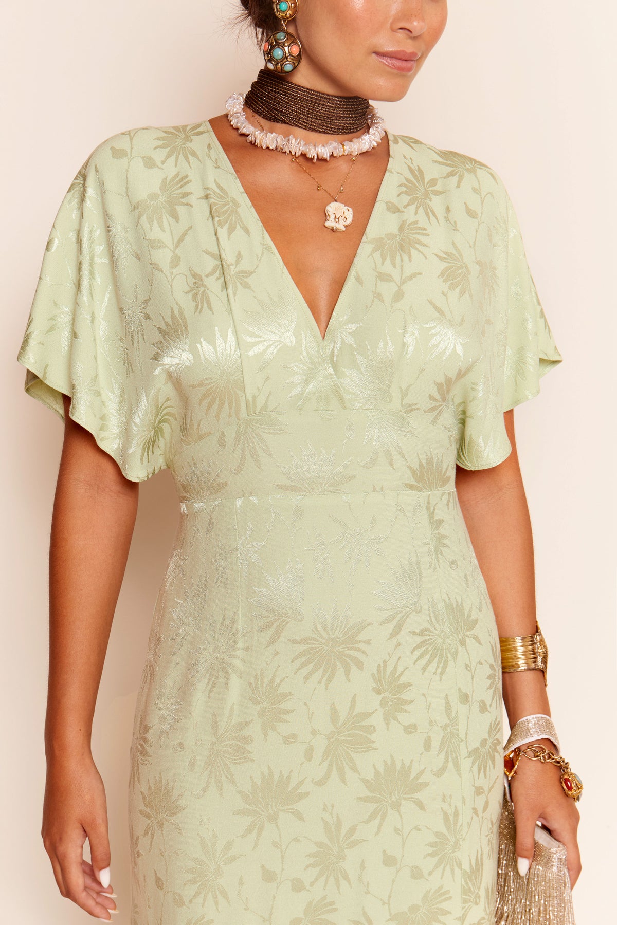 A woman wears the Sadie - Daisy Jacquard Sage dress, a light green, 1930s-inspired piece with short sleeves and a V-neckline. She accessorizes with layered necklaces, bangles, statement earrings, and holds a gold fringed clutch. Her face is partly out of frame.