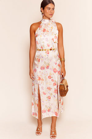 A woman wears the Valerie - Waterblossom Ivory sleeveless halter dress with a front slit, styled with a gold belt, sandals, and gold jewelry. She carries a woven handbag and has her hair pulled back.