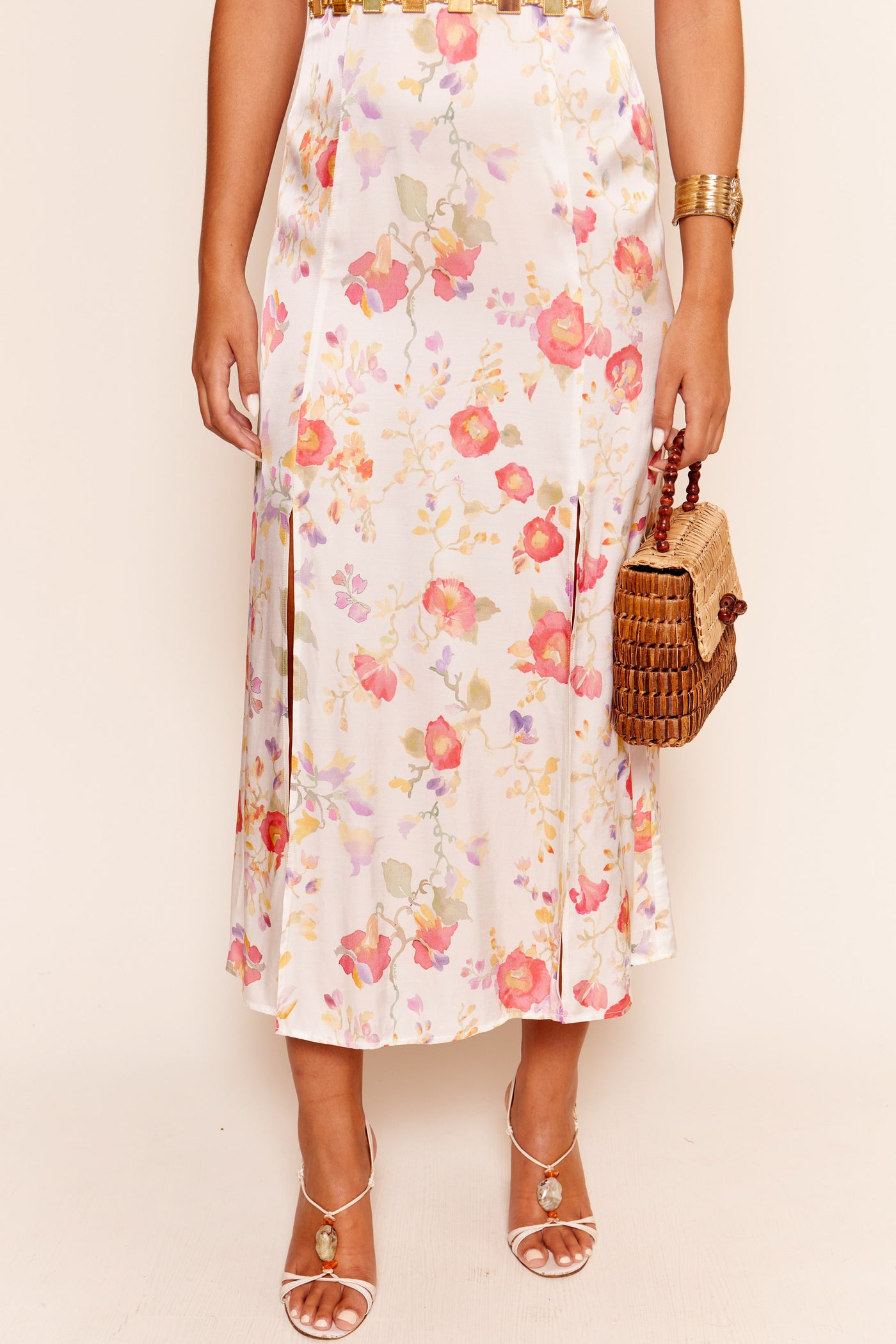 Wearing the Valerie - Waterblossom Ivory midi skirt, white strappy heels, a gold cuff bracelet, and holding a woven rattan handbag. Their upper body is not visible.