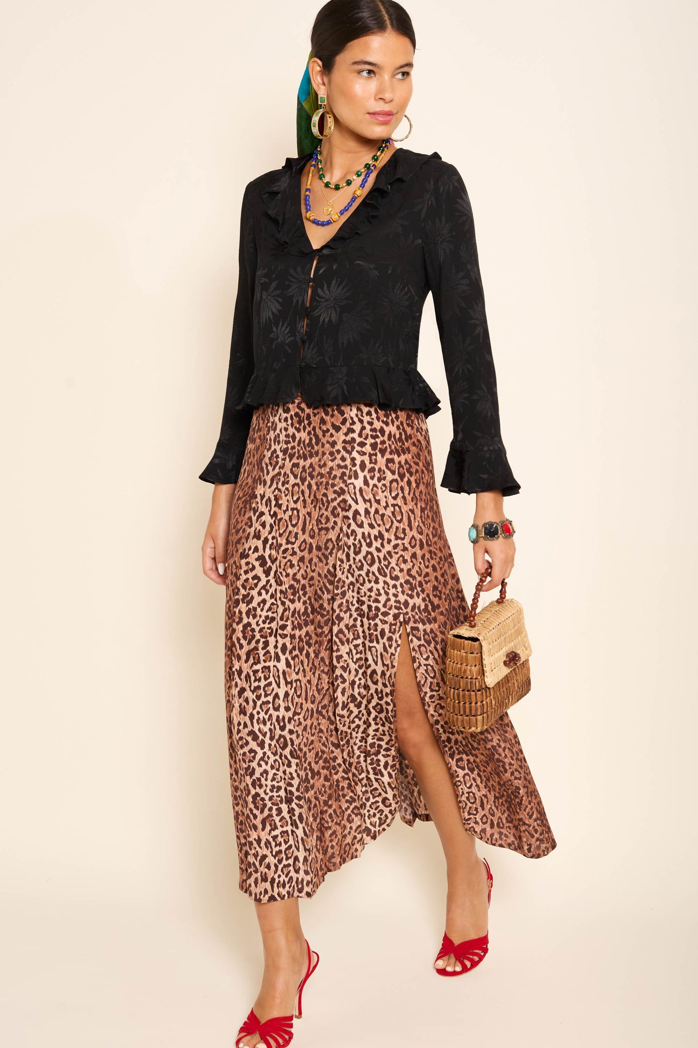 A woman stands against a light background, wearing a black blouse, the Georgia - Leopard silk midi skirt with a front slit, red strappy heels, colorful jewelry, and carrying a woven handbag.