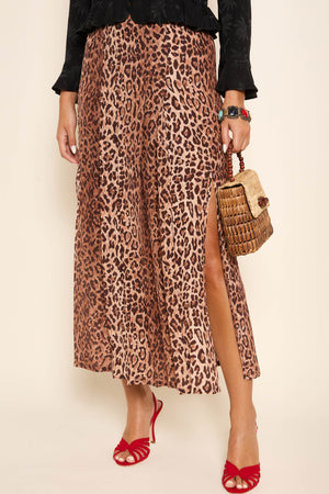 A person wearing the Georgia - Leopard skirt with a side slit, paired with red heeled sandals, a woven handbag, and a black top. Only the lower half of their body is visible.
