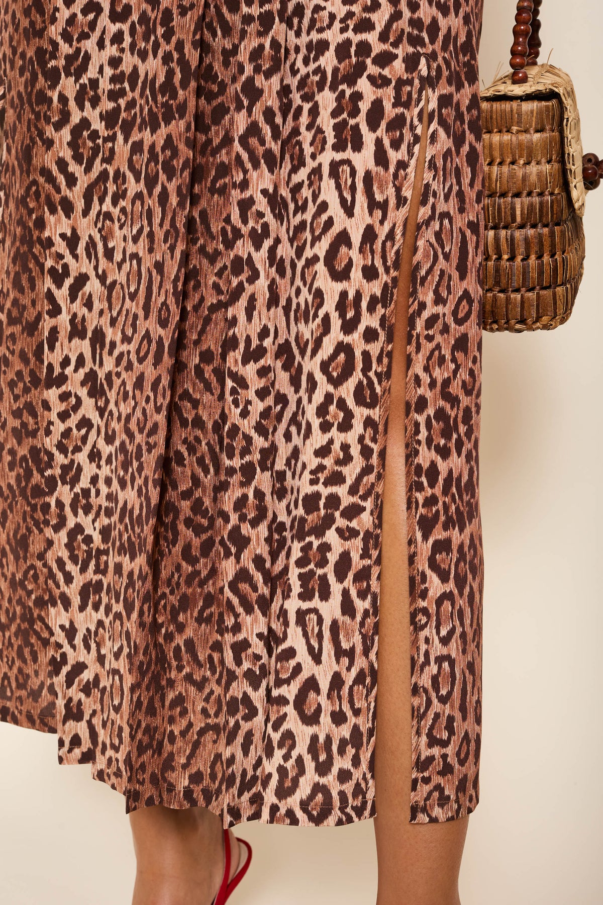 A close-up of a person wearing the Georgia - Leopard silk midi skirt with a side slit, holding a woven brown handbag. Their leg is slightly visible through the slit, and they are paired with red shoes.
