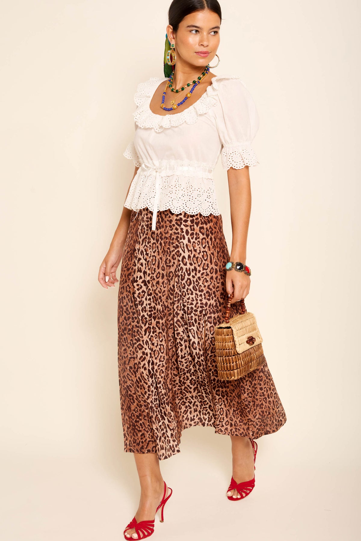 A woman wears a white lace blouse, the Georgia - Leopard silk midi skirt, red open-toe heels, colorful jewelry, and carries a woven handbag while posing against a neutral background.