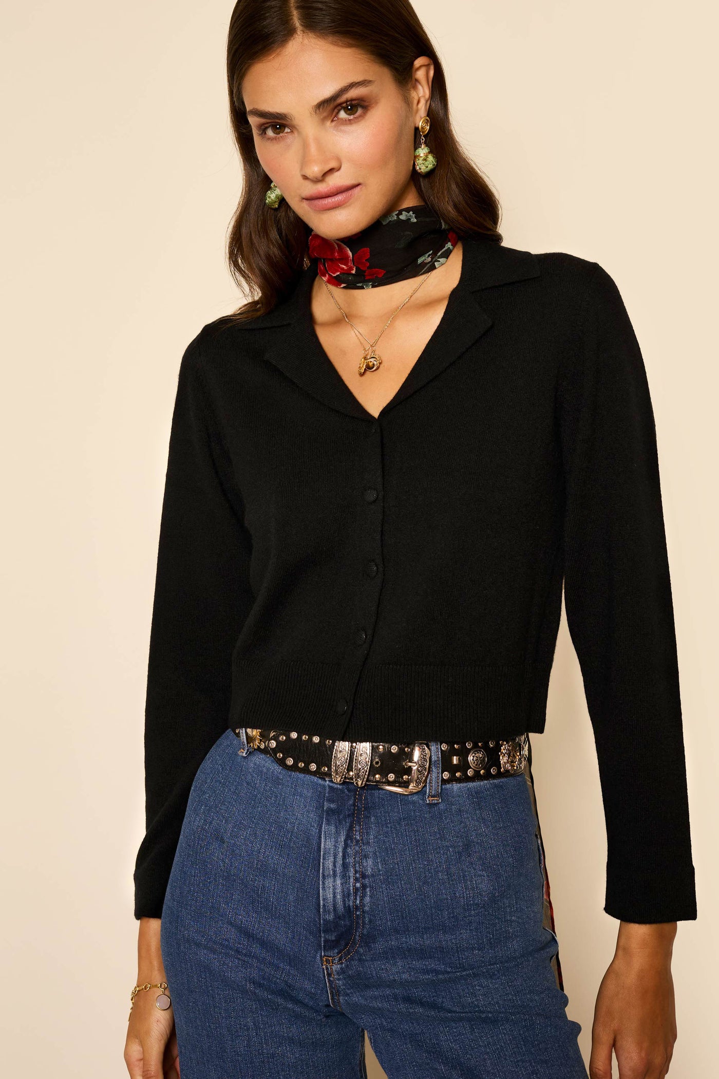 A woman wearing the Carla - Black cardigan, blue jeans, a black belt with silver details, and a black scarf with red floral print. She has long brown hair, earrings, and layered necklaces, standing against a beige background.