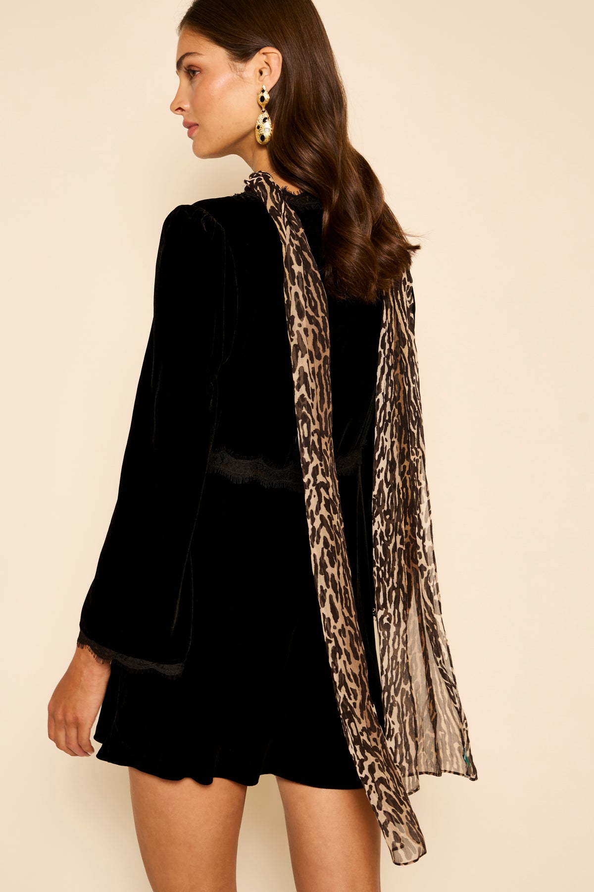 A woman stands against a neutral background wearing a black long-sleeve dress, gold statement earrings, and the Julien - Bohemia Leopard sheer leopard print scarf draped over her shoulder.