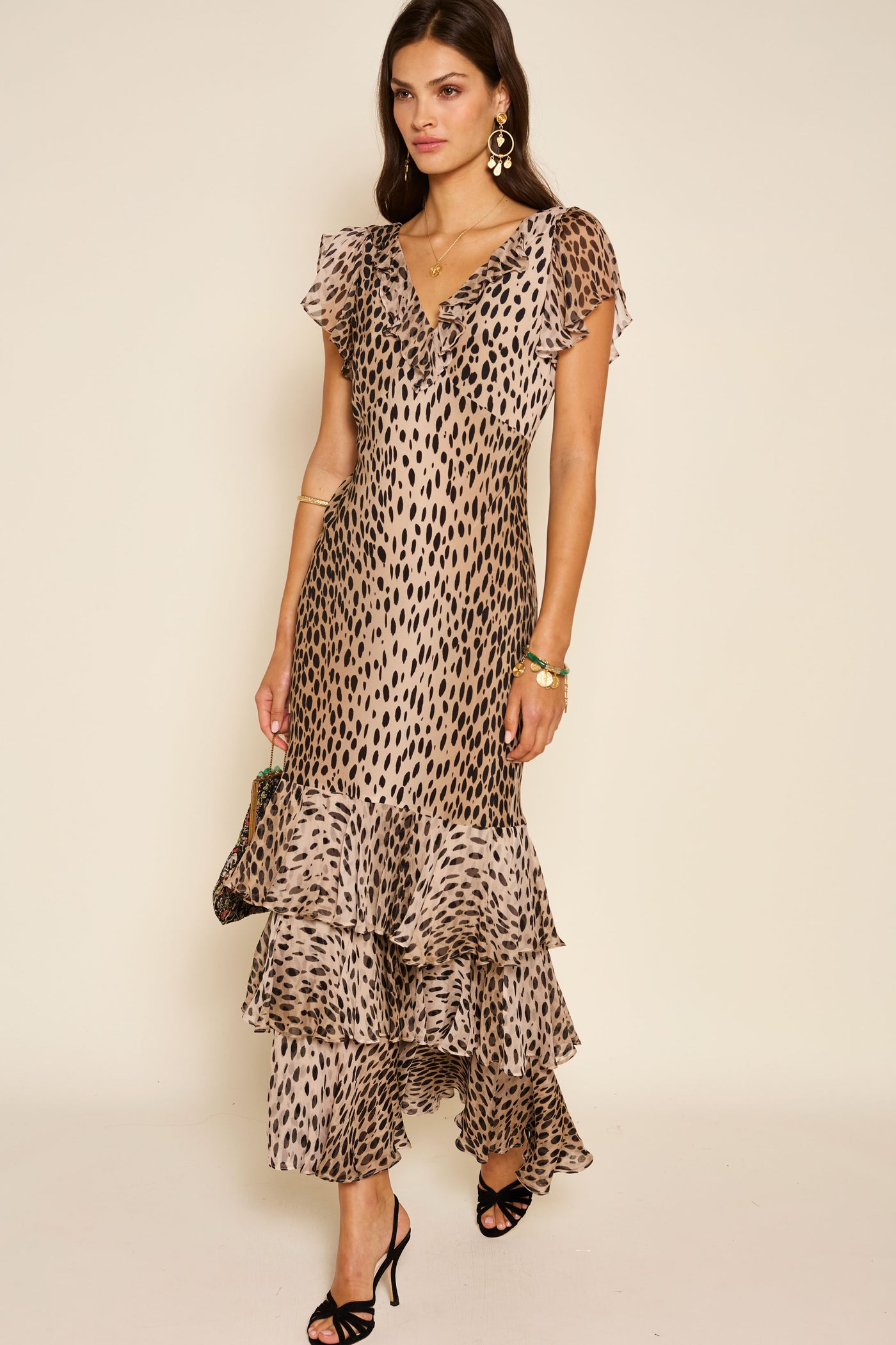 A woman in the Amelle - Antelope Spot Black ankle-length silk crepe de chine dress with a beige and black animal print, gold earrings, and black heels stands against a plain light background, holding a small bag at her side.