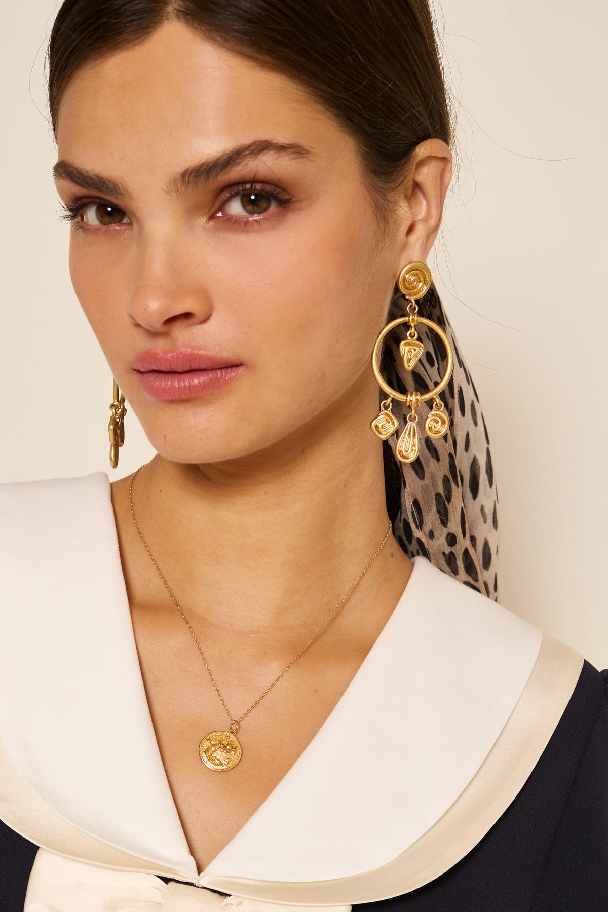 A woman with brown hair wears a cream-collared outfit, gold earrings, and the Zodiac - Aquarius elegant gold vermeil necklace. She has a neutral expression, with a spotted scarf tied in her hair and draped over her shoulder.