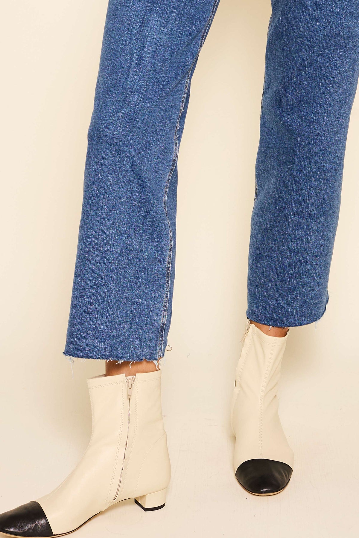 A person wearing blue denim jeans with raw hems and dainty Rey - Ivory two-tone leather ankle boots with black toe caps and side zippers, standing on a light-colored floor.