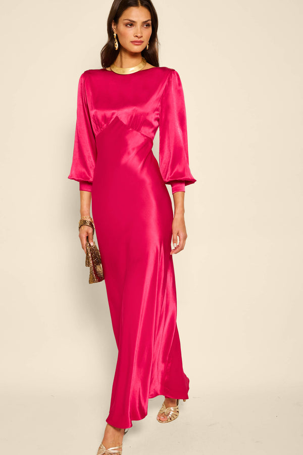 A woman models the marni - raspberry gown with a vintage silhouette, three-quarter sleeves, and fitted waist, accessorized with a small gold bag, gold earrings, and sandals against a plain light background.