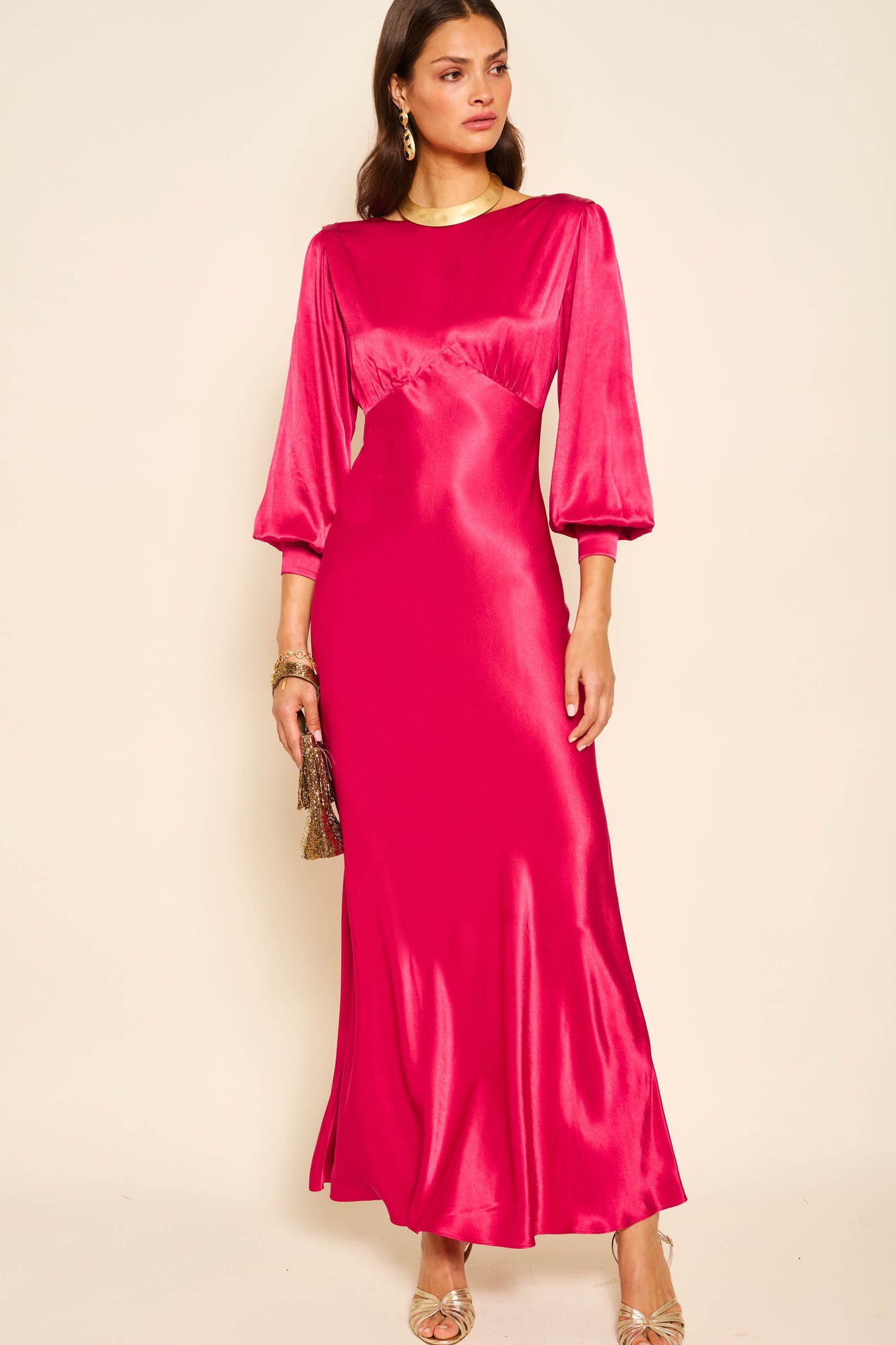 A woman in a long marni - raspberry gown with a vintage-inspired silhouette, gold earrings and bracelet, metallic heels, and a small handbag stands against a plain light background.