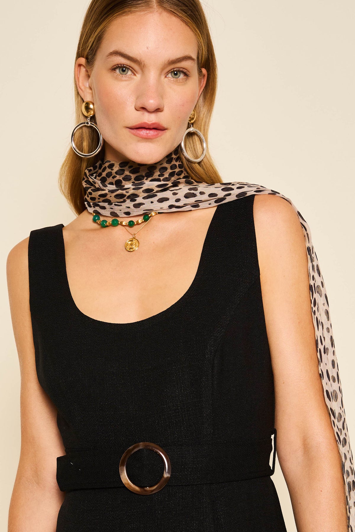 A woman wears a sleeveless black dress with a belt, gold hoop earrings, a green beaded necklace, and the Julien Scarf Antelope Spot Black in sheer leopard-print georgette draped over one shoulder.