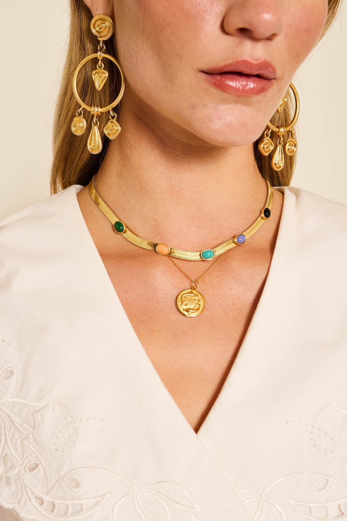 A woman in a cream embroidered top wears large gold vermeil charm earrings and the Zodiac - Pisces necklace, layered with colorful gemstones and a round pendant.