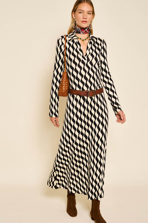 A woman stands against a plain beige background wearing the Tillie - Mono Wave long-sleeve jersey knit dress, brown belt, brown boots, a brown shoulder bag, and a colorful scarf.