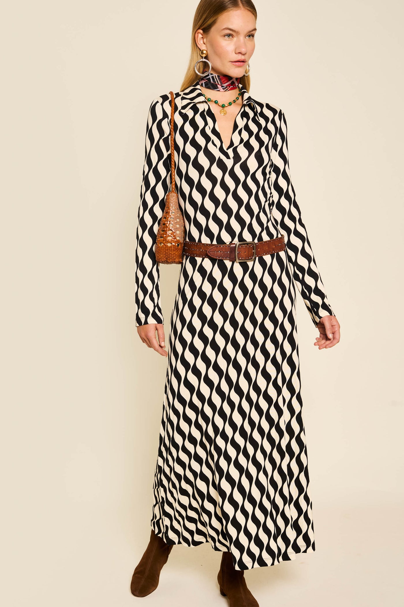 Wearing the Tillie - Mono Wave long-sleeve dress, a woman pairs it with a brown belt, boots, shoulder bag, colorful earrings, and a patterned scarf against a light background.