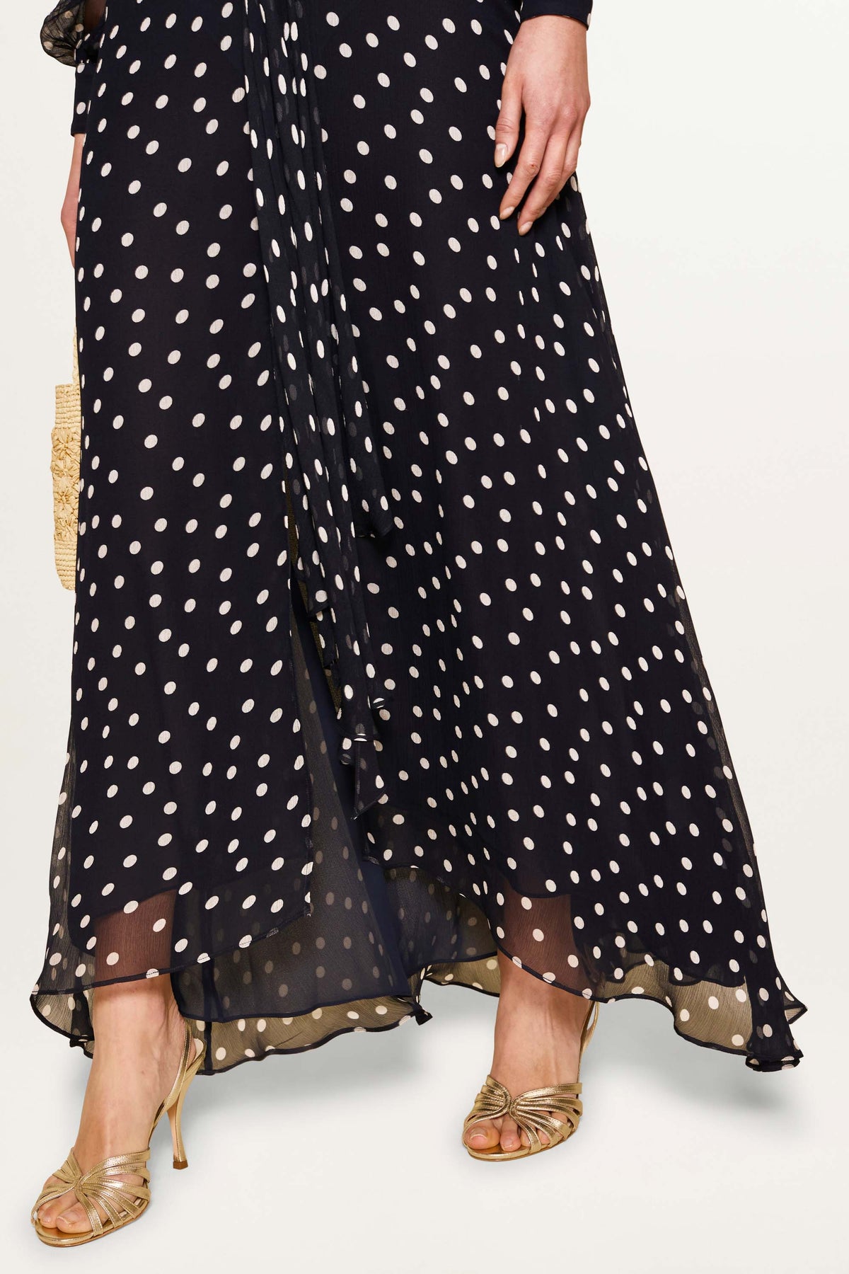 A person in a Meera - Bohemia Spot Deep Navy gown with white polka dots and long crinkled-chiffon sleeves stands against a light background, wearing gold strappy heels and holding a small woven bag.