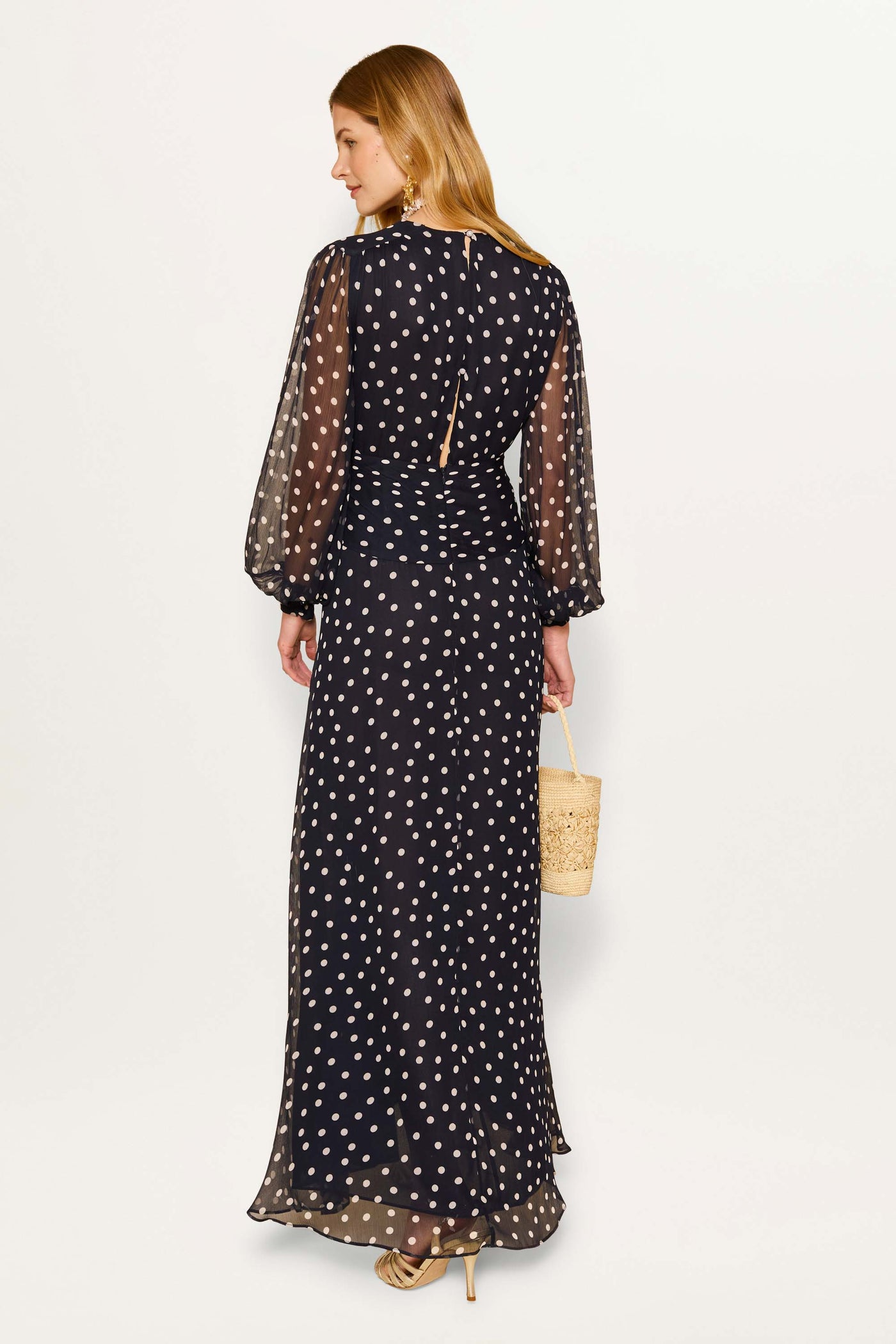 A woman with long hair wears the Meera - Bohemia Spot Deep Navy gown, featuring white polka dots and sheer long sleeves. She pairs it with beige heels and a straw handbag, standing against a plain white background.
