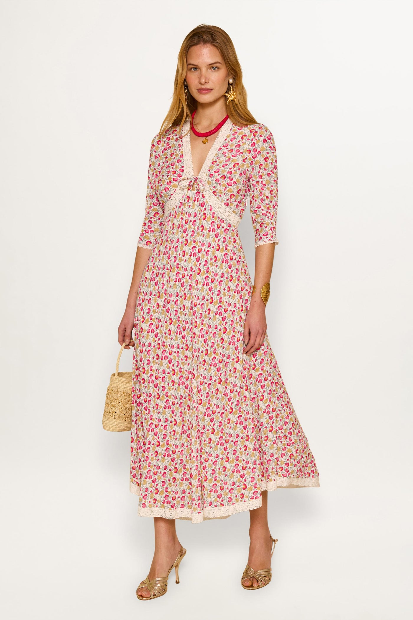 A woman poses in the Amina - Shell Pansy Ivory dress, featuring a fitted bodice and flared skirt. She accessorizes with a woven handbag, heeled sandals, gold jewelry, and wavy blonde hair against a plain white background.