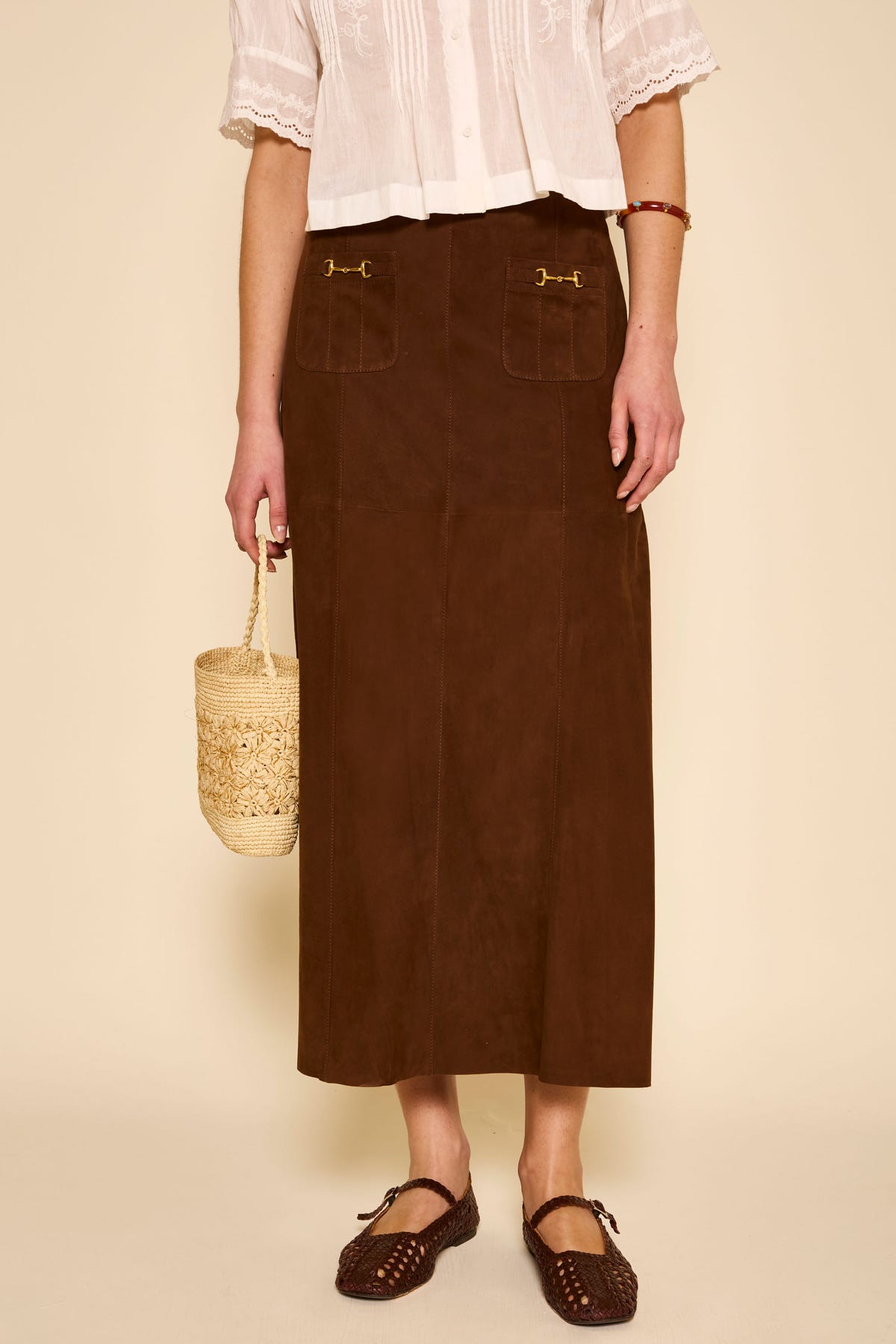 A person wearing a white short-sleeve blouse and a long vintage-inspired skirt with front pockets stands holding a woven basket bag. The outfit is completed with the Edwina - Brown woven shoes. The person is photographed from shoulders down.