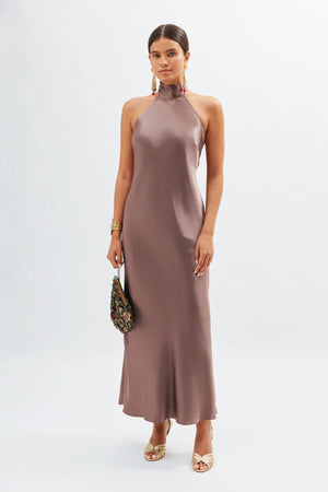 A woman poses against a white background wearing the Maura - Mink, a sleeveless satin halterneck gown with an open back. She holds a small patterned handbag, gold strappy heels, statement earrings, and a bracelet.