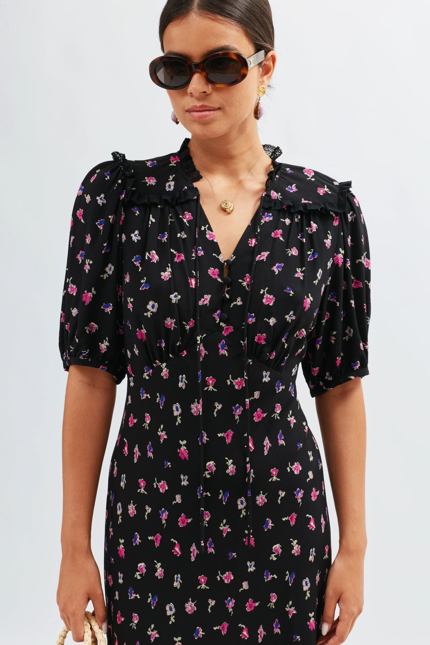 A woman in the Gwen – French Ditsy Black dress, featuring delicate floral prints, puffed short sleeves, and ruffle details, stands against a light backdrop. Styled with dark sunglasses and gold jewelry—perfect for day-to-night looks.