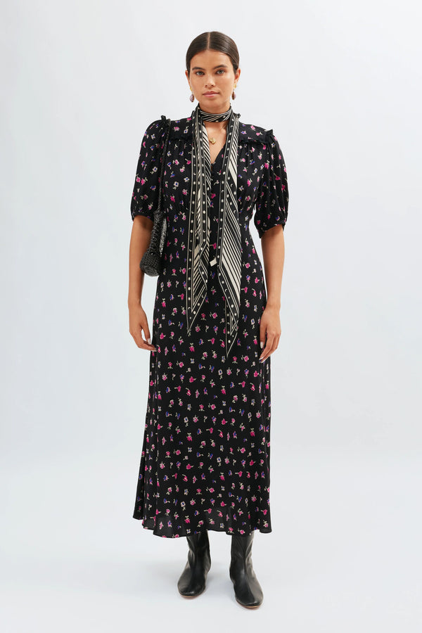 A woman stands against a plain background wearing the Gwen - French Ditsy Black dress, an ankle-length black piece with pink and white florals. She styles it with black ankle boots, a patterned scarf, and a black purse; her hair is pulled back.