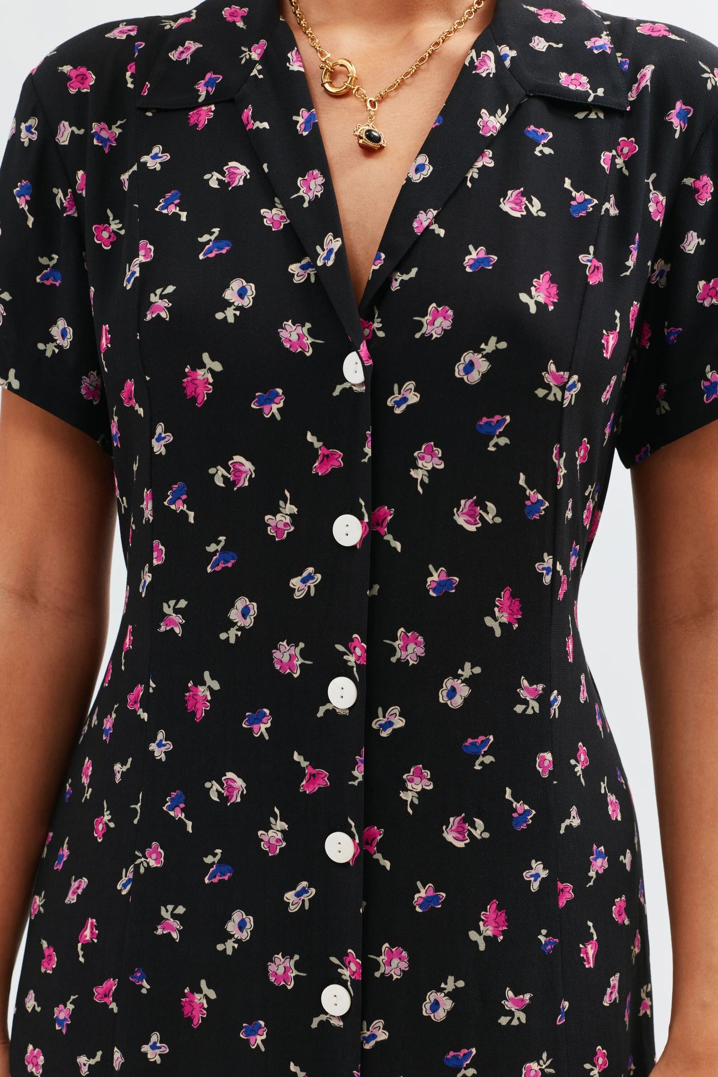 A close-up of a person wearing the Florrie - French Ditsy Black, a vintage-inspired black button-down tea dress with pink, purple, and white floral ditsy print, styled with a gold necklace featuring black stones.
