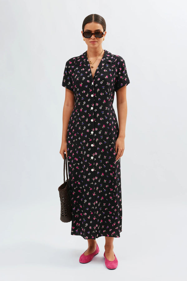A woman wearing the Florrie - French Ditsy Black tea dress stands with a black woven bag, pink flats, gold necklace, and sunglasses.
