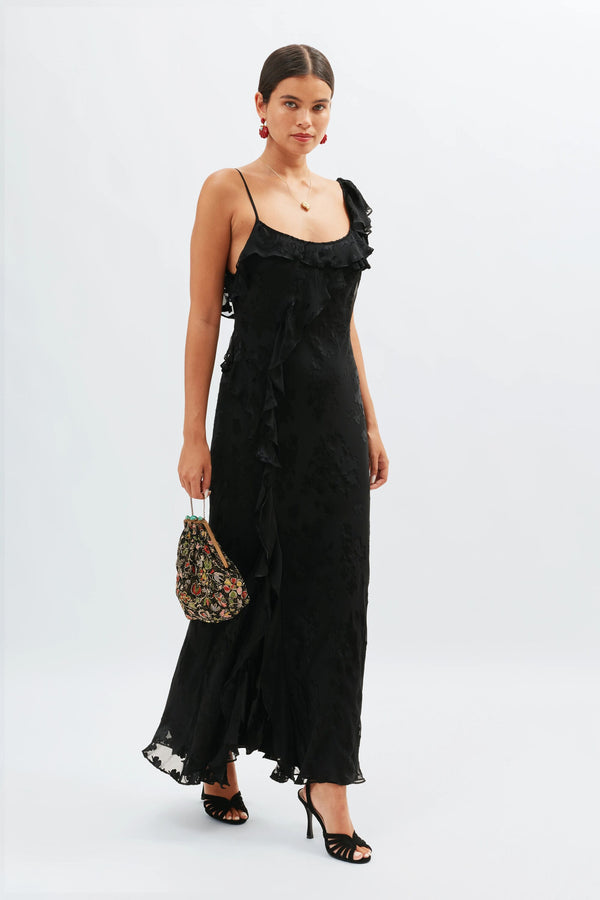 A woman wearing the Ester - Bouquet Devore Black long ruffled dress with thin straps stands holding a small floral handbag. She has her hair tied back, wears black heels, and accessorizes with earrings and a necklace against a plain background.