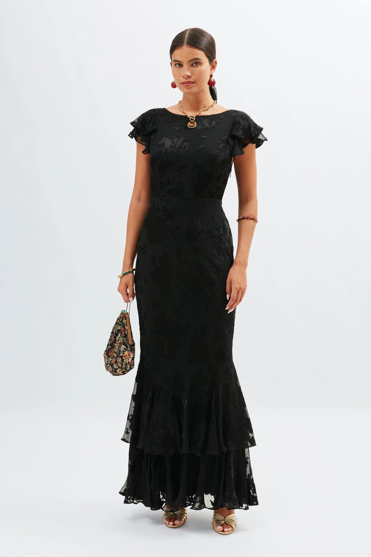 A woman in the fitted, floor-length Ordinia - Bouquet Devore Black gown with ruffled sleeves and hem stands against a white background, holding a patterned handbag. Gold jewelry and sandals complete her black-tie wedding look.