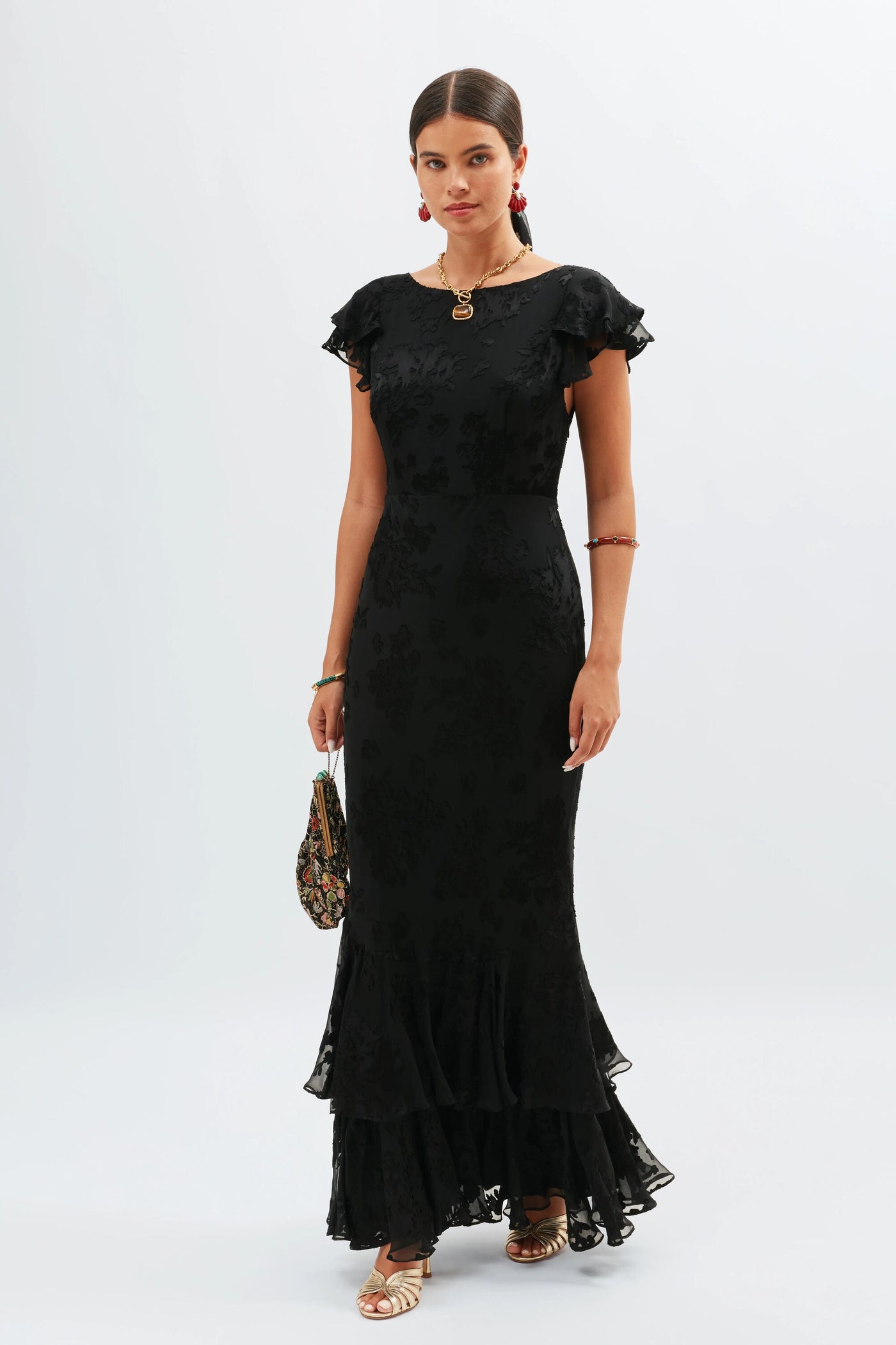 A woman wears the fitted, 1930s-inspired Ordinia - Bouquet Devoré Black gown with short ruffled sleeves and a flared hem, gold sandals, statement necklace and earrings, and carries a small patterned handbag against a plain light background.