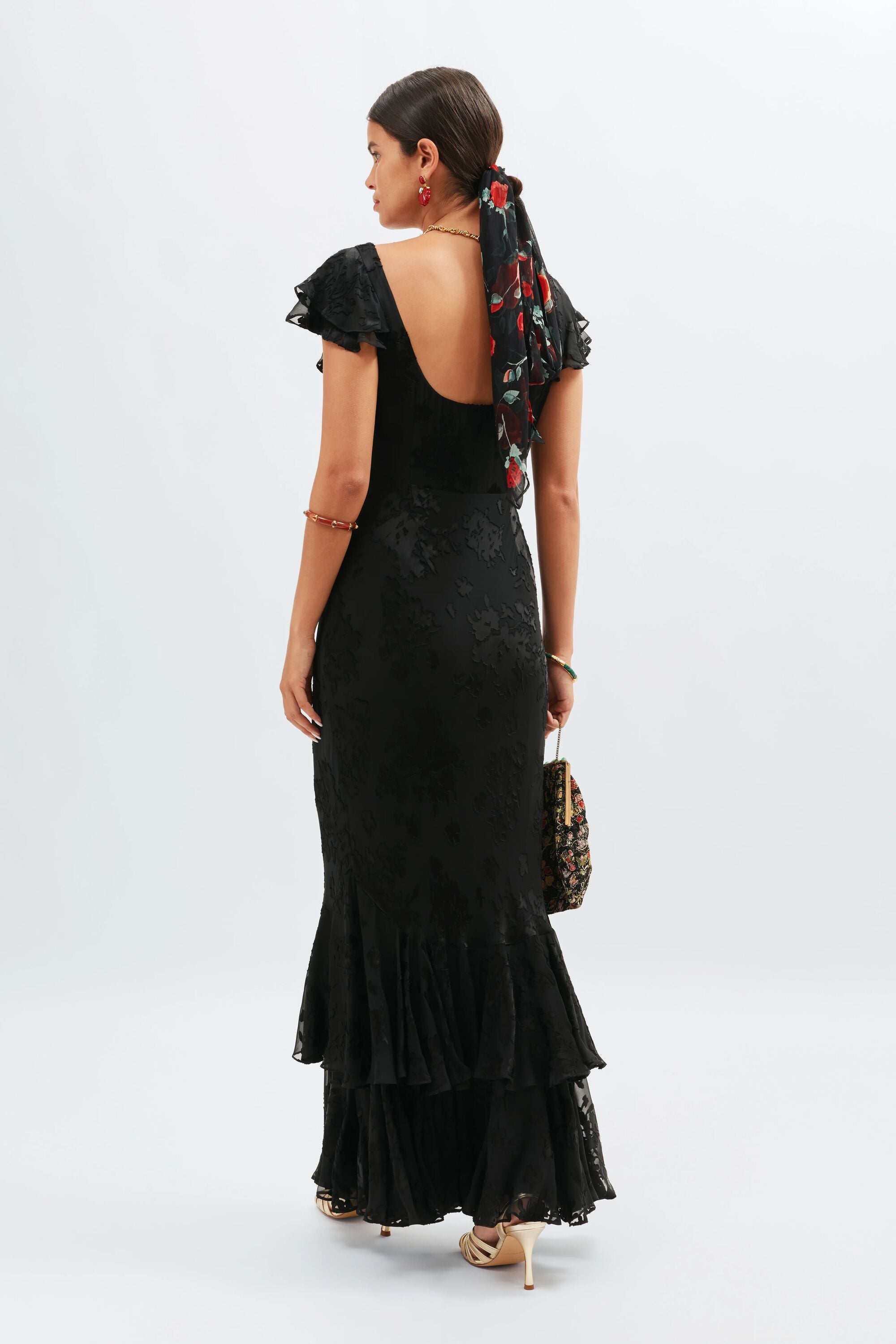 A woman faces away, wearing the 1930s-inspired Ordinia - Bouquet Devoré Black gown with flutter sleeves and a ruffled hem. She accessorizes with a floral hair scarf, gold hoop earrings, and a patterned handbag—ideal for black-tie weddings.