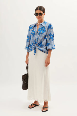 A woman wearing sunglasses, the Blake - Milos Floral Blue shirt tied at the waist, a long white skirt, black flip-flops, and carrying a black woven bag stands against a plain white background.