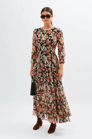 A woman in the Kristen - Primrose Floral Deep Navy dress, styled with brown ankle boots, dark sunglasses, and a black handbag, stands against a plain white backdrop.