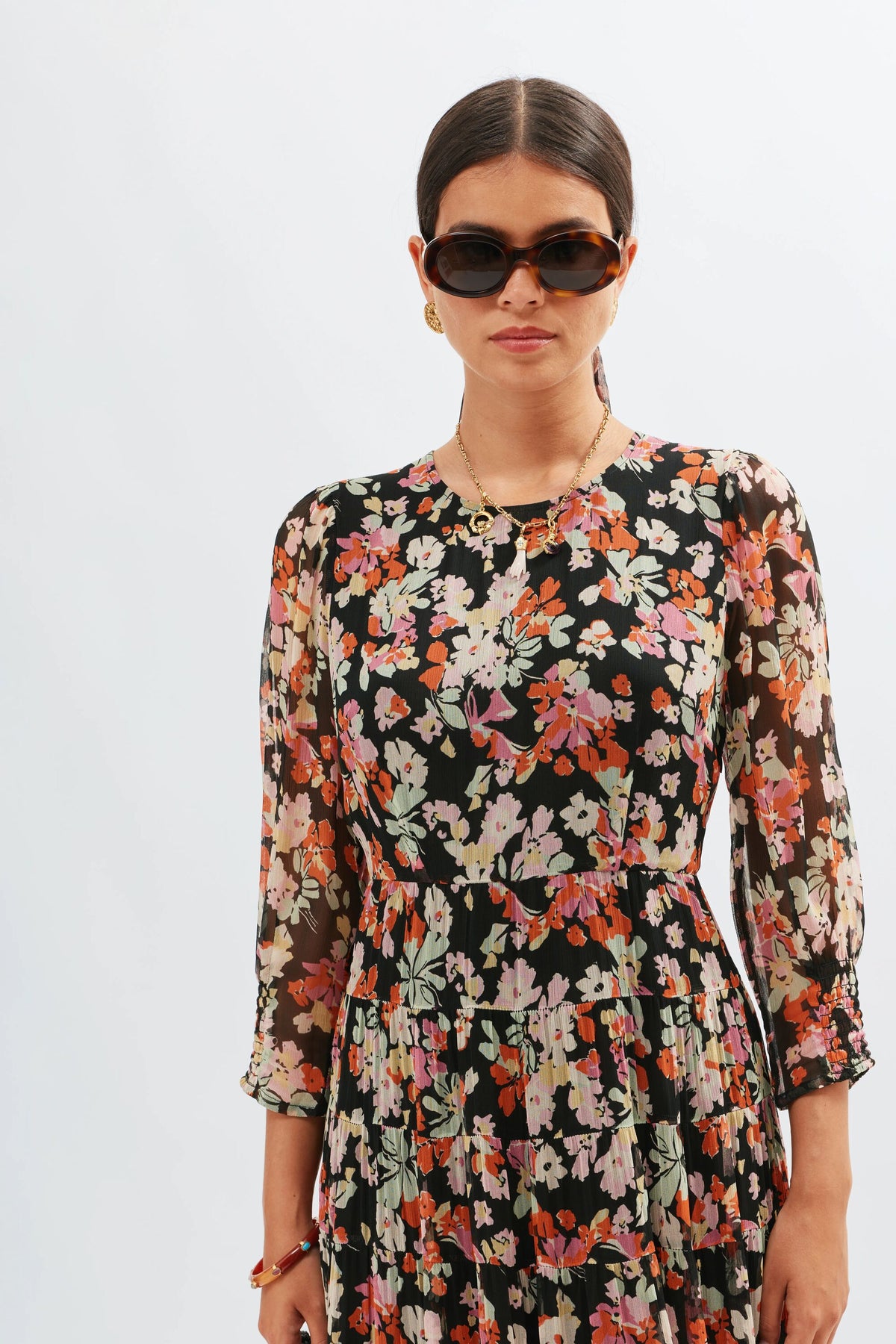 A woman in dark sunglasses and earrings poses against a white background, wearing the Kristen - Primrose Floral Deep Navy dress with vintage-inspired prints and chic ankle boots. She has shoulder-length dark hair and accessorizes with a necklace.