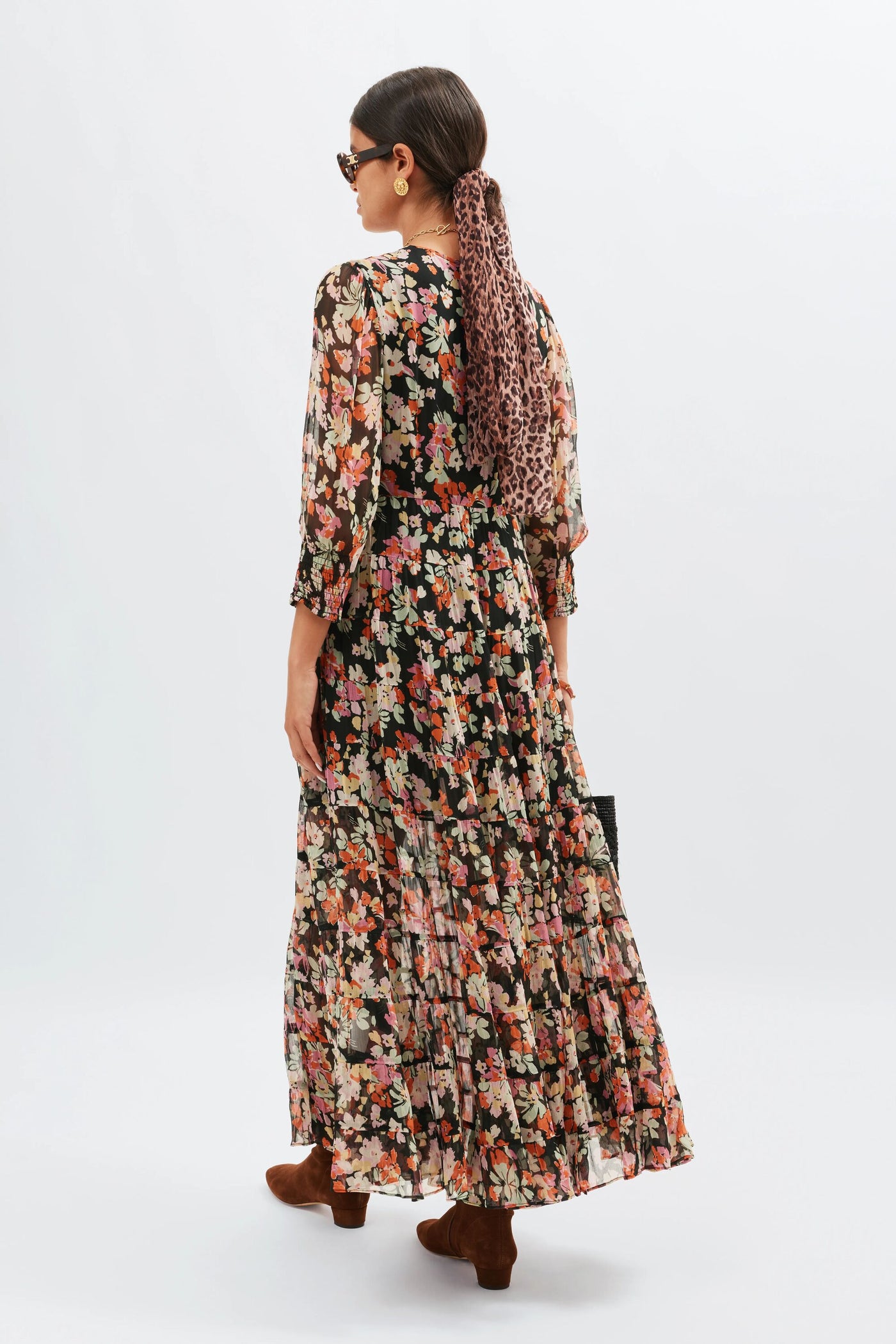 Woman in the long Kristen - Primrose Floral Deep Navy dress with tiered layers, brown ankle boots, sunglasses, and gold earrings. Her hair is tied back with a leopard-print scarf as she carries a black handbag and faces away from the camera.