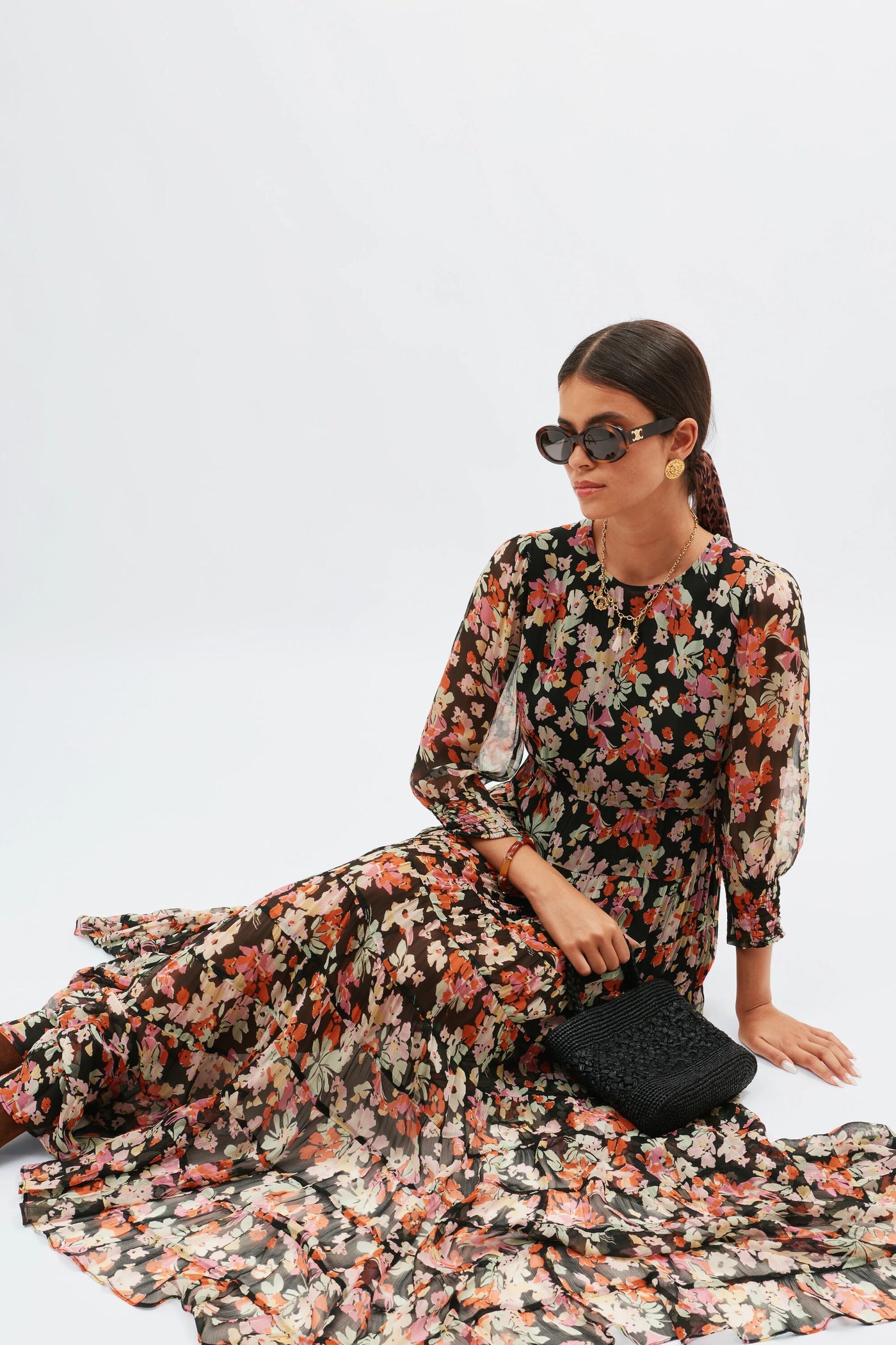 A woman in large sunglasses and the Kristen - Primrose Floral Deep Navy dress sits on the floor with a black textured clutch, set against a plain white background.