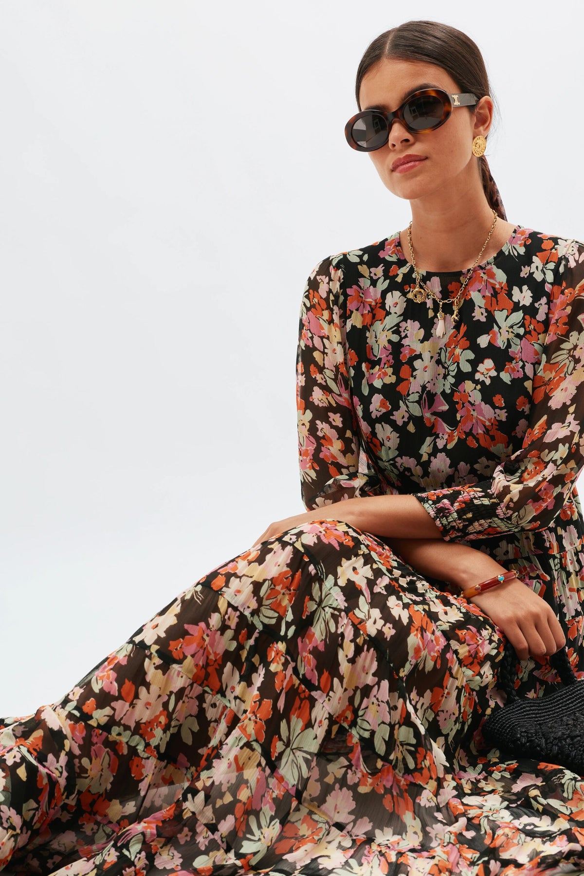A woman wears large sunglasses and the Kristen - Primrose Floral Deep Navy dress, featuring vintage-inspired floral prints. She sits against a white background with her hair pulled back, accessorized with gold earrings and a necklace.