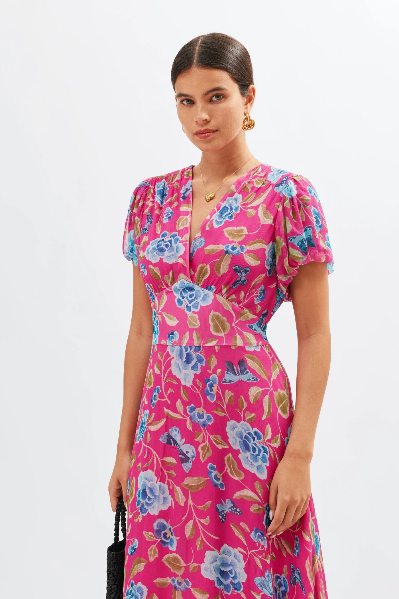 A woman stands against a white background wearing the Corrine - Butterfly Floral Magenta dress featuring blouson sleeves, a V-neckline, and bold blue and purple patterns. She pairs it with gold earrings, a necklace, and a black bag.