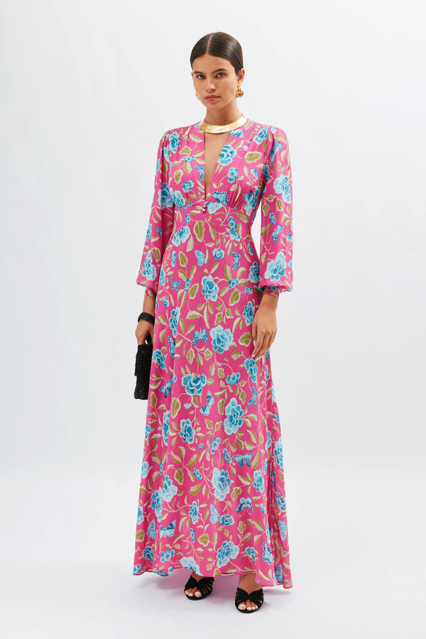 A woman wears the Emory - Butterfly Floral Magenta dress, featuring a long silhouette with blue and green floral patterns, long sleeves, and a deep V-neck. She accessorizes with a black clutch, black sandals, and her hair is slicked back.