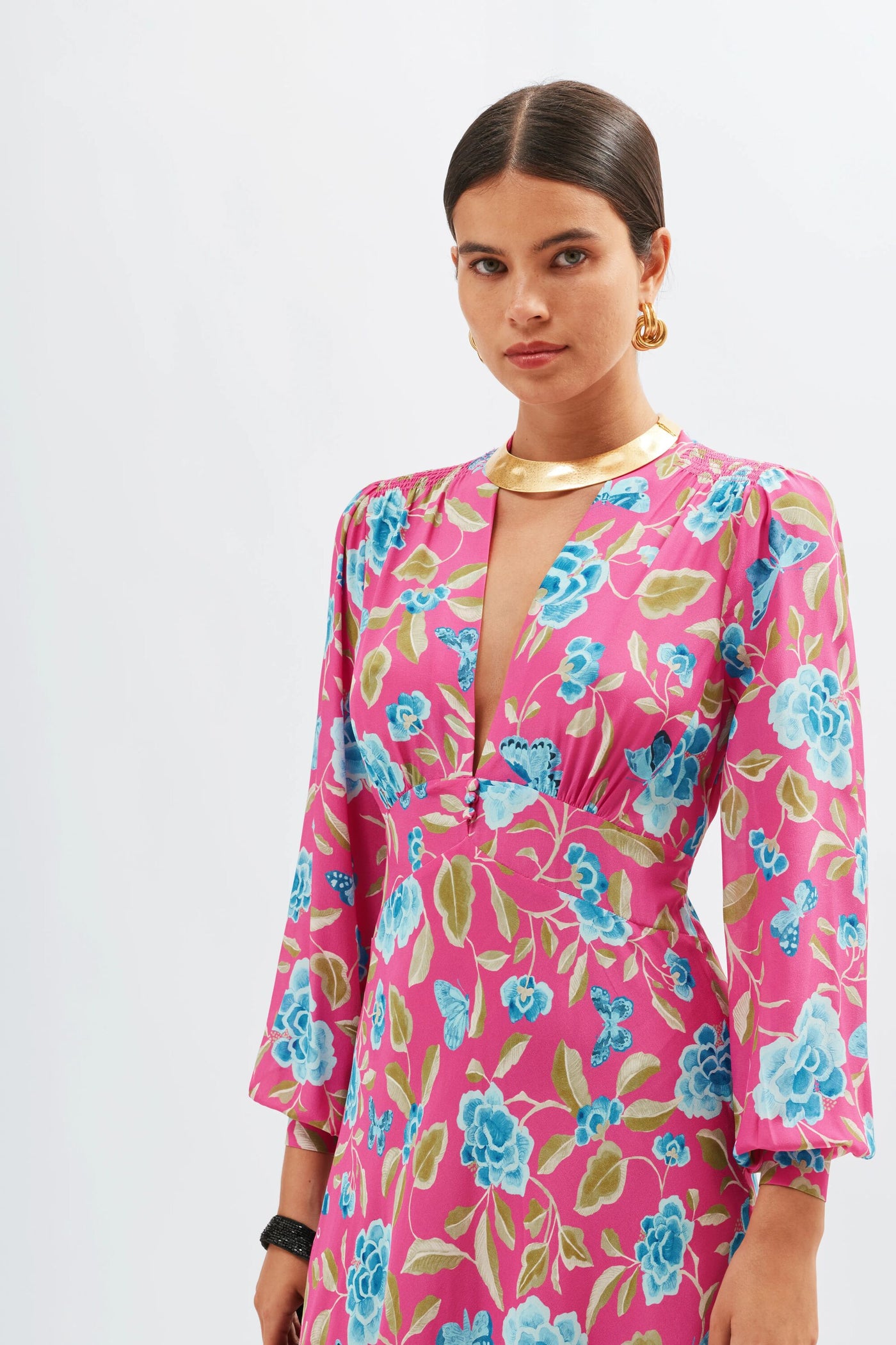 A woman wears the Emory - Butterfly Floral Magenta long-sleeve dress, gold hoop earrings, and a gold choker necklace, with her dark hair parted in the middle, standing against a plain white background.