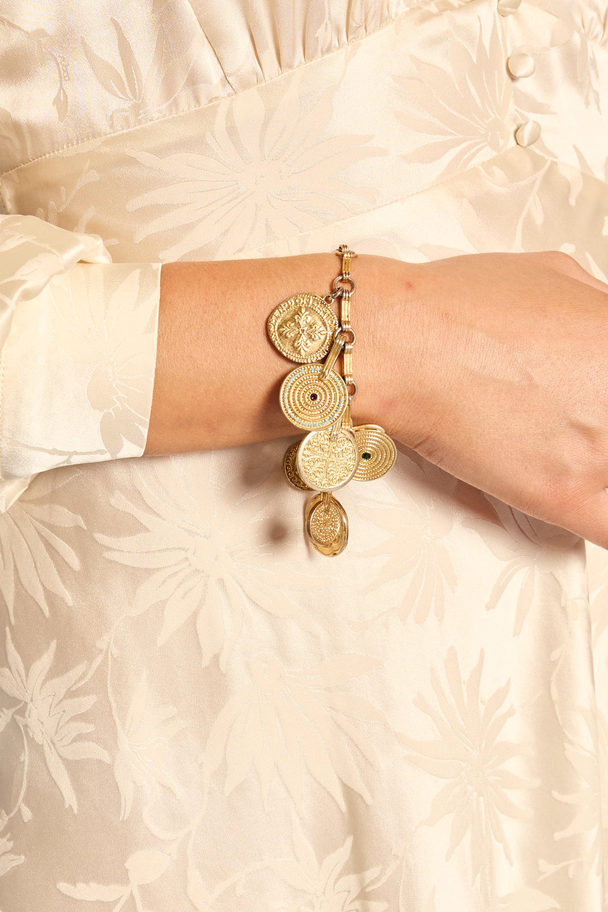 Wearing a cream floral satin dress with fabric buttons, a person showcases the Adie - Gold bracelet, a bohemian accessory adorned with golden charms, on their wrist.