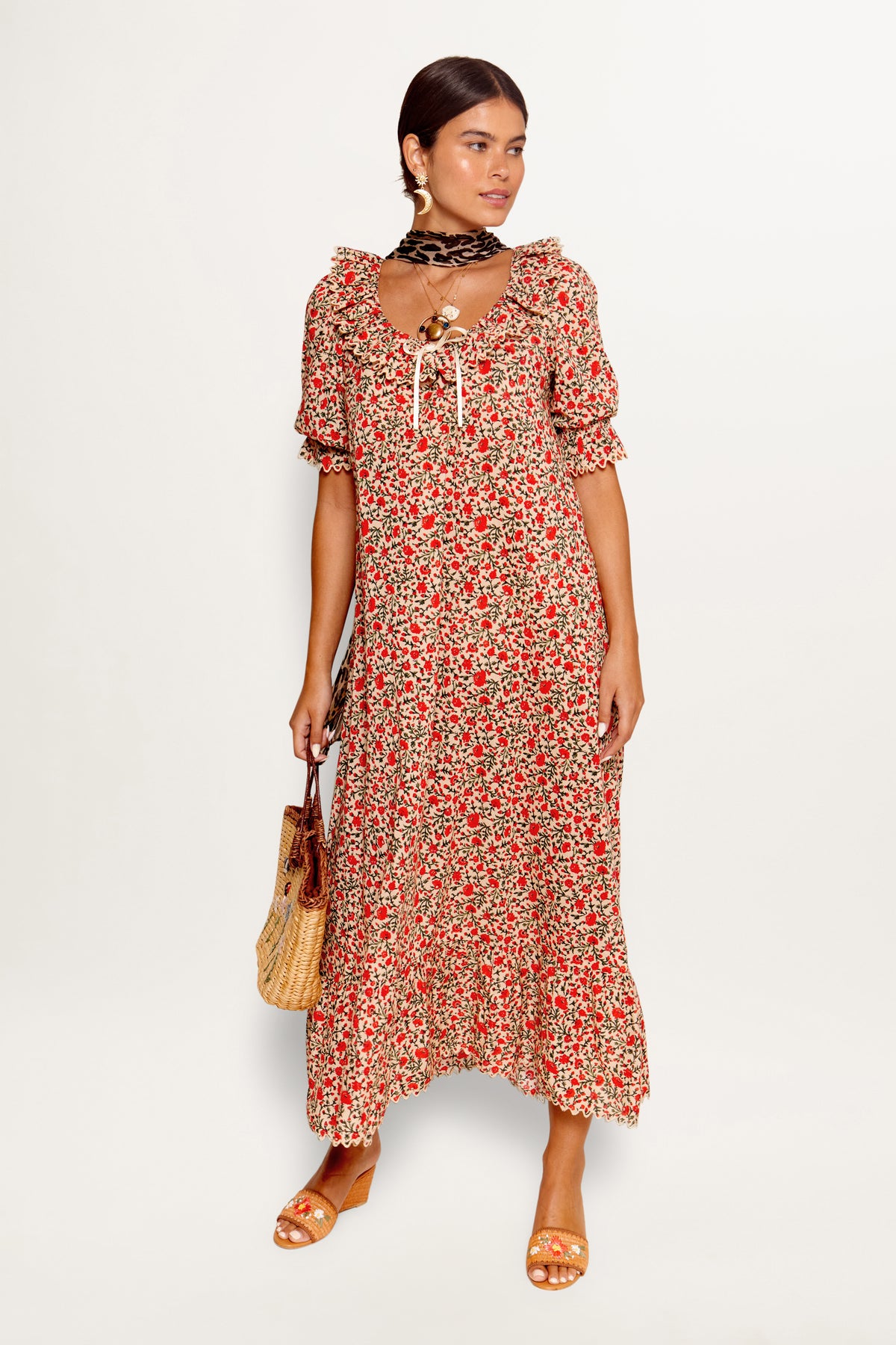 A woman wears the Aprilia - Ditsy Trail Cream maxi dress with puff sleeves, tan sandals, a wicker handbag, and a black patterned scarf around her neck, standing against a white background.
